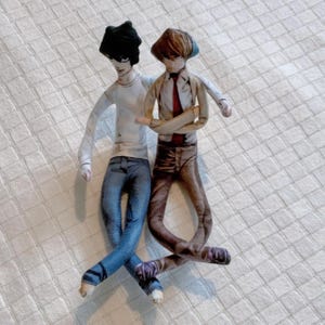 May include: Two handmade dolls, one in a white shirt and blue jeans, the other in a tan shirt, brown trousers, and a red tie. The dolls are posed together, with one arm around the other. The dolls have detailed facial features and are on a white quilted surface.
