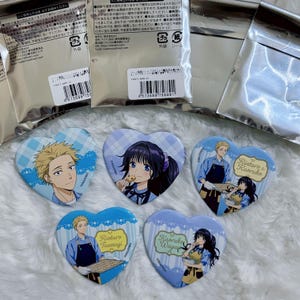 May include: Heart-shaped anime character badges with various designs. One features a blonde character, while others depict a girl eating a cookie and a couple baking. The badges have a blue and white color scheme.