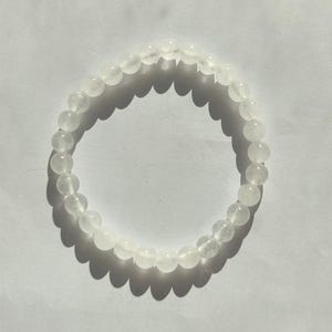 Könnte beinhalten: Ein weißes Perlenarmband. Das Armband besteht aus kleinen, runden, durchscheinenden Perlen, die aneinandergereiht sind. Das Armband liegt auf einer weißen Oberfläche, mit Schatten, die von den Perlen geworfen werden.