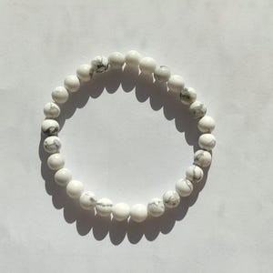 Könnte beinhalten: Weißes Perlenarmband mit grauer Marmorierung. Das Armband besteht aus runden, polierten Perlen. Die Perlen sind zu einer kreisförmigen Form aufgereiht. Das Armband liegt auf einer weißen Oberfläche.