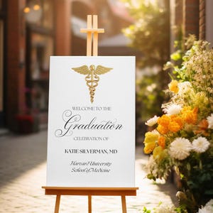 Op de afbeelding: Een wit bord met een gouden medisch symbool en de tekst "Welkom bij de afstudeerceremonie van Katie Silverman, MD Harvard University School of Medicine" op een ezel.