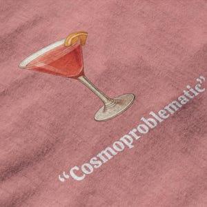 May include: A pink fabric with a watercolor illustration of a cocktail in a stemmed glass, garnished with an orange slice. The text "Cosmoproblematic" is printed in white.