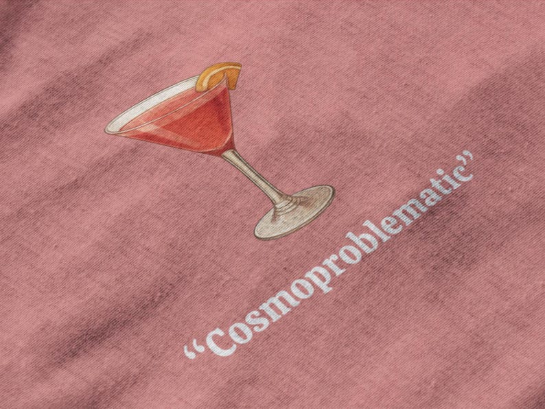 May include: A pink fabric with a watercolor illustration of a cocktail in a stemmed glass, garnished with an orange slice. The text "Cosmoproblematic" is printed in white.
