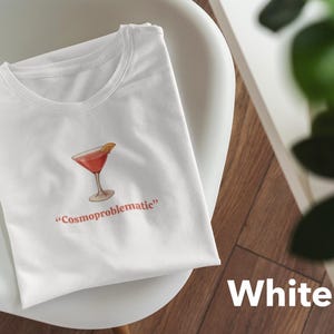 May include: White t-shirt with a cocktail graphic and the text "Cosmoproblematic" in red. The cocktail is a red drink with a lemon slice. The shirt is on a white chair with a wooden background.
