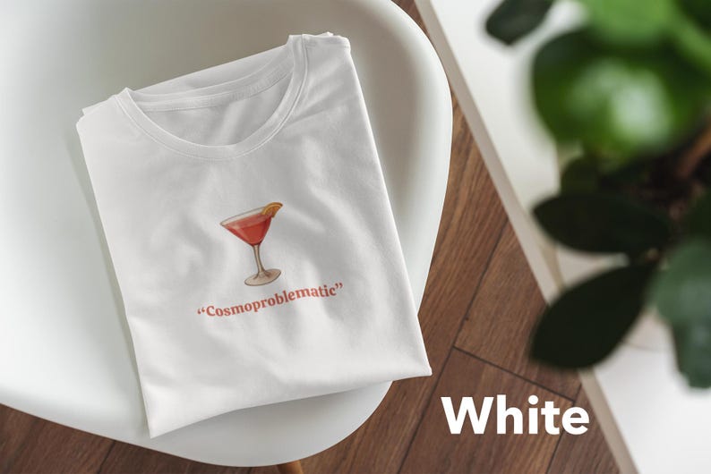 May include: White t-shirt with a cocktail graphic and the text "Cosmoproblematic" in red. The cocktail is a red drink with a lemon slice. The shirt is on a white chair with a wooden background.