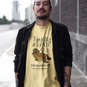 May include: A light yellow t-shirt with the text "Top it like it's HOT" and a cartoon hot dog wearing sunglasses and a hat. Below the graphic is "THE GLIZZY LAB" and the tagline "Lab tested. Street approved."