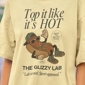 May include: A light yellow t-shirt with the text "Top it like it's HOT" and "THE GLIZZY LAB". The shirt features a cartoon hot dog wearing sunglasses and a cap. The graphic includes the text "Relish The Moment Errday" and "Lab tested. Street approved."