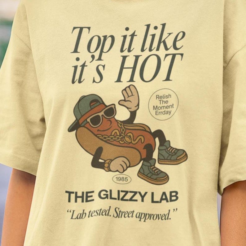 May include: A light yellow t-shirt with the text "Top it like it's HOT" and "THE GLIZZY LAB". The shirt features a cartoon hot dog wearing sunglasses and a cap. The graphic includes the text "Relish The Moment Errday" and "Lab tested. Street approved."