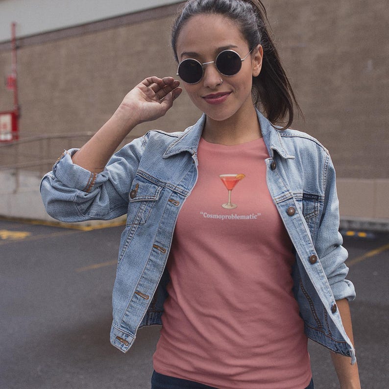 May include: A dusty rose t-shirt featuring a cocktail graphic with the text "Cosmoproblematic" in white. The shirt is worn with a light blue denim jacket and sunglasses.