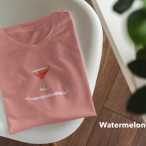 May include: A watermelon-colored t-shirt with a cocktail graphic and the text "Cosmoproblematic." The shirt is folded and resting on a white chair. A plant is visible in the background.