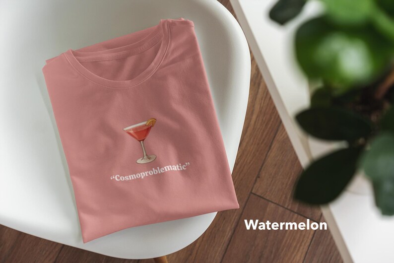 May include: A watermelon-colored t-shirt with a cocktail graphic and the text "Cosmoproblematic." The shirt is folded and resting on a white chair. A plant is visible in the background.