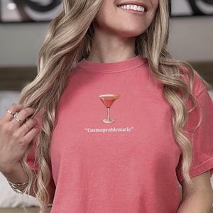 May include: A coral-colored t-shirt featuring a cocktail glass graphic with a lemon slice. Below the graphic, the text reads "Cosmoproblematic". The shirt has a crew neck and short sleeves.