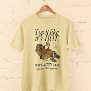 May include: A light yellow t-shirt with a graphic of a hot dog wearing sunglasses and a hat. The text "Top it like it's HOT" and "THE GLIZZY LAB" are printed on the shirt. The shirt is hanging on a wooden hanger.