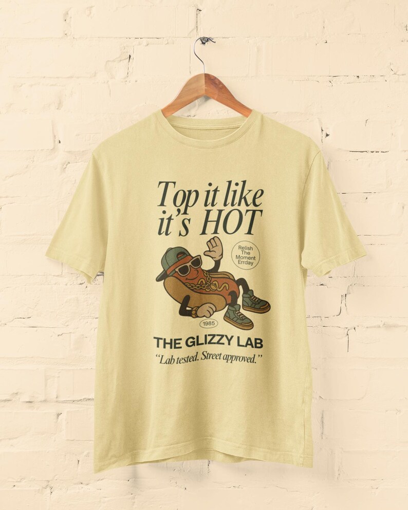 May include: A light yellow t-shirt with a graphic of a hot dog wearing sunglasses and a hat. The text "Top it like it's HOT" and "THE GLIZZY LAB" are printed on the shirt. The shirt is hanging on a wooden hanger.