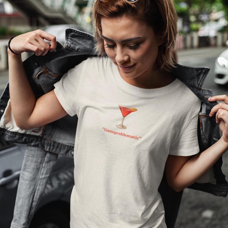 May include: Off-white t-shirt featuring a watercolor illustration of a cocktail with a lemon garnish. The text "Cosmoproblematic" is printed below the image in red. The shirt is made of a soft, light-colored fabric.