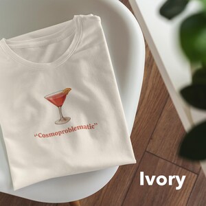 May include: Ivory-colored t-shirt featuring a watercolor illustration of a cocktail in a martini glass with an orange slice. The text "Cosmoproblematic" is printed below the image in red. The shirt is folded on a white chair.