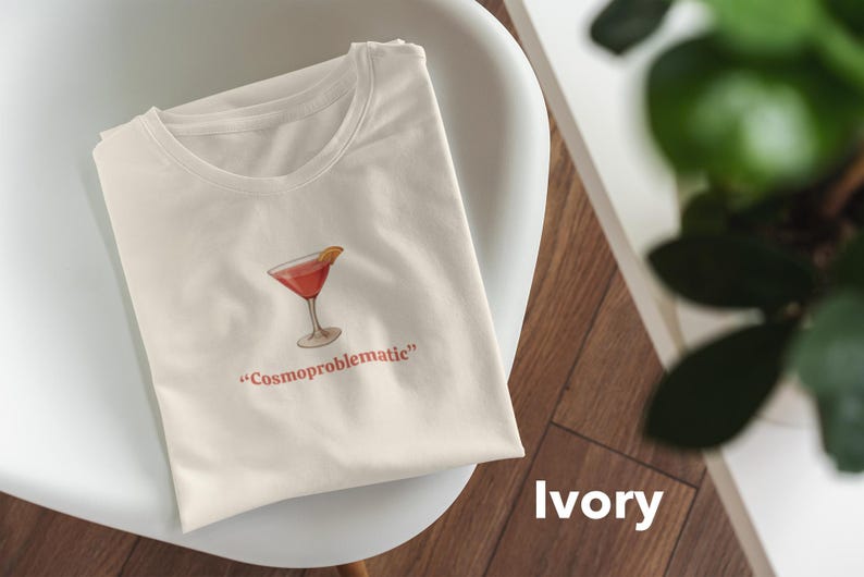 May include: Ivory-colored t-shirt featuring a watercolor illustration of a cocktail in a martini glass with an orange slice. The text "Cosmoproblematic" is printed below the image in red. The shirt is folded on a white chair.
