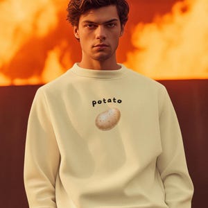 Pode incluir: Camisola creme com a palavra "potato" impressa em preto acima de uma ilustração realista de uma batata. A camisola tem gola redonda e mangas compridas.
