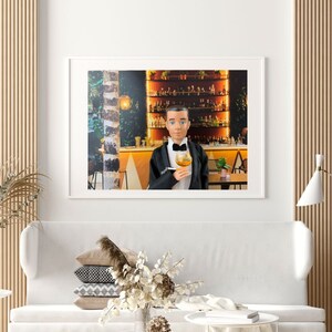 May include: A framed photograph of a man in a tuxedo holding a glass of amber liquid. He is standing in front of a bar with shelves of bottles behind him. The background is a warm, inviting color.