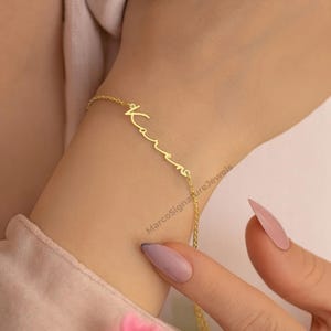 Puede incluir: Una delicada pulsera dorada con el nombre "Karen" escrito en cursiva. La pulsera se lleva en la muñeca, y la cadena es fina y elegante. La imagen está bien iluminada, resaltando los detalles de la joya. El nombre de la marca "MarcoSignatureJewels" es visible.
