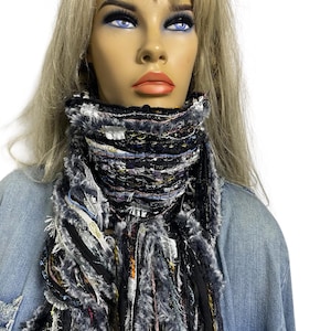 May include: A textured scarf with fringing in black, grey, and white. The scarf features a layered design with a thick, woven collar and long, flowing fringe. The scarf is worn over a denim jacket.