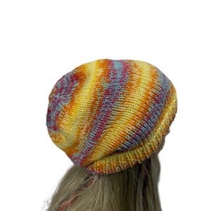 May include: A colorful knitted beanie hat with a yellow, orange, pink, and blue striped pattern. The hat is worn on a person with long blonde hair.