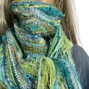 Puede incluir: Bufanda multicolor hecha a mano con un diseño texturizado y con flecos. La bufanda presenta tonos de turquesa, verde y amarillo, con acentos blancos y dorados. Está hecha de varios hilos y telas.