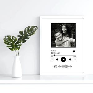 Puede incluir: Marco blanco con foto en blanco y negro de una pareja. Debajo, un gráfico de reproductor de música muestra la canción "Perfect" de Ed Sheeran. Un jarrón blanco con hojas verdes a la izquierda.