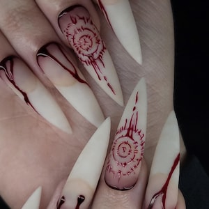 May include: Long, white, stiletto-shaped acrylic nails with a red, dripping blood effect. Some nails have a red, circular design with a symbol in the center.