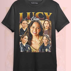 May include: Black t-shirt featuring a graphic of Lucy Chen with the text "LUCY Chen" in gold lettering. The design includes multiple images of the actress. The shirt is made of a soft material.