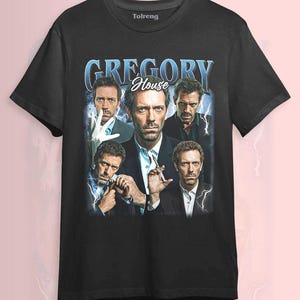 Puede incluir: Camiseta negra con un diseño vintage que presenta múltiples imágenes de Gregory House y el texto "GREGORY House" en azul. La camiseta está hecha de un material suave y es adecuada para ropa casual.