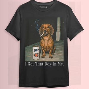 Op de afbeelding: Zwart T-shirt met een bruine teckel met een sigaret in zijn mond en een blikje Lite bier. De tekst "I Got That Dog In Me" staat eronder. Het shirt heeft korte mouwen.