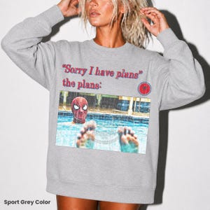 May include: A sport grey crewneck sweatshirt featuring the red text "Sorry I have plans" and "the plans:" above a rectangular graphic of a person wearing a red and blue Spider-Man mask in a swimming pool with feet visible in the foreground.