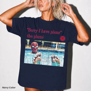 May include: A navy blue short-sleeve t-shirt featuring the red text "Sorry I have plans" and "the plans:" above a graphic of a person wearing a Spider-Man mask in a swimming pool, with a small red and blue circular spider logo on the right side.