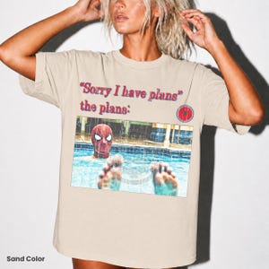 May include: A sand-colored t-shirt featuring the text "Sorry I have plans" and "the plans:" in red and blue lettering. Below the text is a graphic of a person wearing a red Spider-Man mask in a swimming pool with feet visible in the foreground.