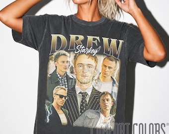 May include: A charcoal gray short-sleeve t-shirt featuring a graphic print of Drew Starkey in various outfits. The text DREW STARKEY is printed in a large, bold font at the top. The design includes lightning bolt accents and a collage of portraits.