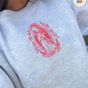 May include: A light gray sweatshirt featuring a red embroidered design of a religious figure within a floral frame. The sweatshirt has a crew neck and long sleeves. The brand logo is visible in the upper right corner.