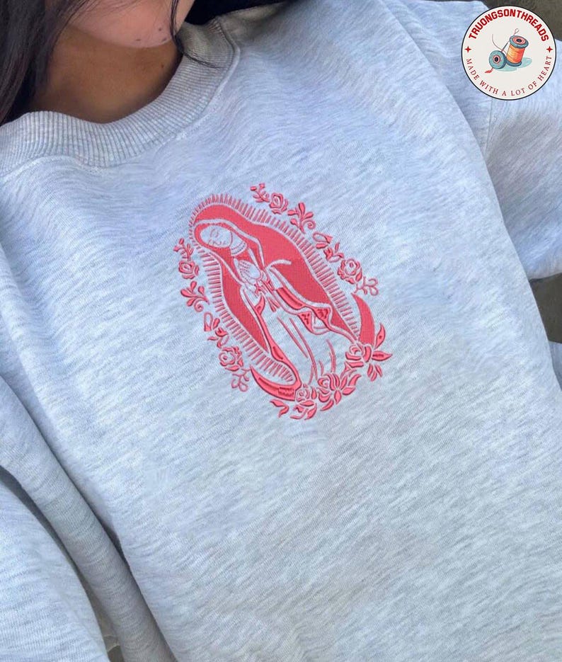 May include: A light gray sweatshirt featuring a red embroidered design of a religious figure within a floral frame. The sweatshirt has a crew neck and long sleeves. The brand logo is visible in the upper right corner.