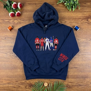 Könnte beinhalten: Ein marineblauer Kapuzenpullover mit einer Grafik von fünf Athleten in verschiedenen Sportuniformen auf der Brust. Der Schriftzug "WINDY CITY" ist in Rot auf dem linken Ärmel gedruckt. Der Hoodie hat lange Ärmel und eine Kängurutasche vorne.