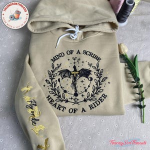May include: Beige hoodie with a hood and white drawstring. The front features an embroidered dragon design with the text "MIND OF A SCRIBE HEART OF A RIDER". The left sleeve has "Fly or die" and flying dragon embroidery.