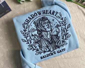 May include: Light blue sweatshirt with an embroidered design featuring a woman with a crown and the text "Shadowheart Baldur's Gate".