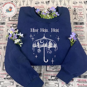 May include: Navy blue sweatshirt with a white graphic of an umbrella, trees, a wolf, and stars. The text "Hoa Hoa Hoa" is above the graphic. The sweatshirt is decorated with purple and white flowers.