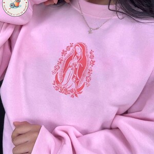 May include: A light pink sweatshirt with a red graphic of a religious figure surrounded by roses. The sweatshirt has long sleeves and a crew neck. The brand logo is visible in the upper left corner.