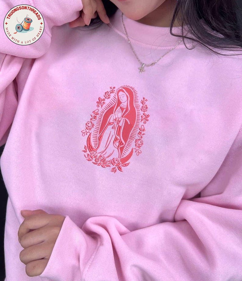 May include: A light pink sweatshirt with a red graphic of a religious figure surrounded by roses. The sweatshirt has long sleeves and a crew neck. The brand logo is visible in the upper left corner.