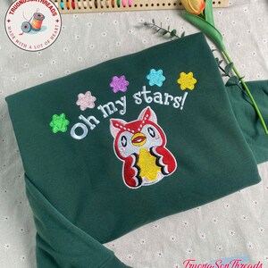 Puede incluir: Una sudadera verde oscuro con un dise&ntilde;o de b&uacute;ho bordado y la frase "Oh my stars!". El b&uacute;ho es rojo, blanco y amarillo, con acentos de estrellas coloridas. La sudadera tiene mangas largas.