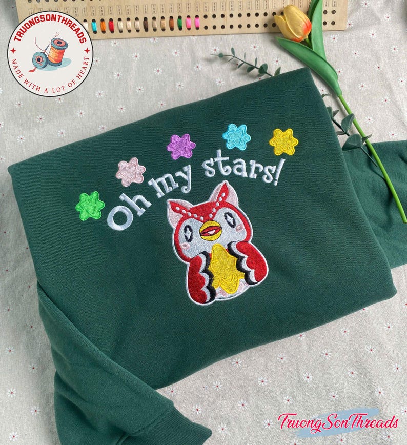 Puede incluir: Una sudadera verde oscuro con un dise&ntilde;o de b&uacute;ho bordado y la frase "Oh my stars!". El b&uacute;ho es rojo, blanco y amarillo, con acentos de estrellas coloridas. La sudadera tiene mangas largas.