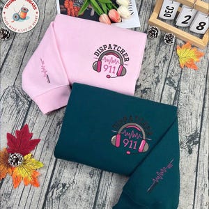 Könnte beinhalten: Zwei Sweatshirts, eines in Rosa und eines in Türkis, mit dem Aufdruck "DISPATCHER" und "911". Das rosa Sweatshirt hat das Design auf der Vorderseite und ein kleineres Design auf dem Ärmel. Das türkisfarbene Sweatshirt hat das Design auf der Vorder- und dem Ärmel.