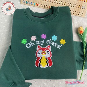 Puede incluir: Una sudadera verde oscuro con la frase bordada "Oh my stars!" sobre un b&uacute;ho de dibujos animados. El b&uacute;ho es rojo, blanco y amarillo con ojos negros. Acentos coloridos en forma de estrella rodean el texto. El logotipo de la marca es visible en la esquina superior izquierda.