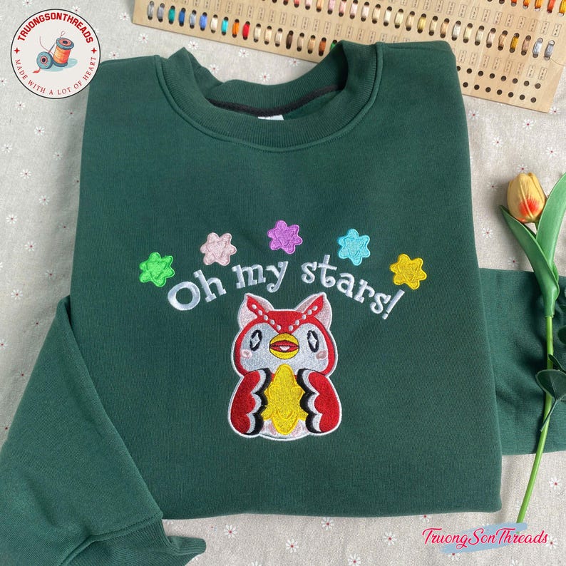 Puede incluir: Una sudadera verde oscuro con la frase bordada "Oh my stars!" sobre un b&uacute;ho de dibujos animados. El b&uacute;ho es rojo, blanco y amarillo con ojos negros. Acentos coloridos en forma de estrella rodean el texto. El logotipo de la marca es visible en la esquina superior izquierda.