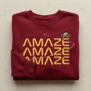 Puede incluir: Una sudadera de manga larga color granate con la palabra AMAZE bordada en hilo dorado tres veces, apilada verticalmente. Sobre el texto hay tres pequeñas estrellas doradas y a la derecha una criatura dorada tipo araña.